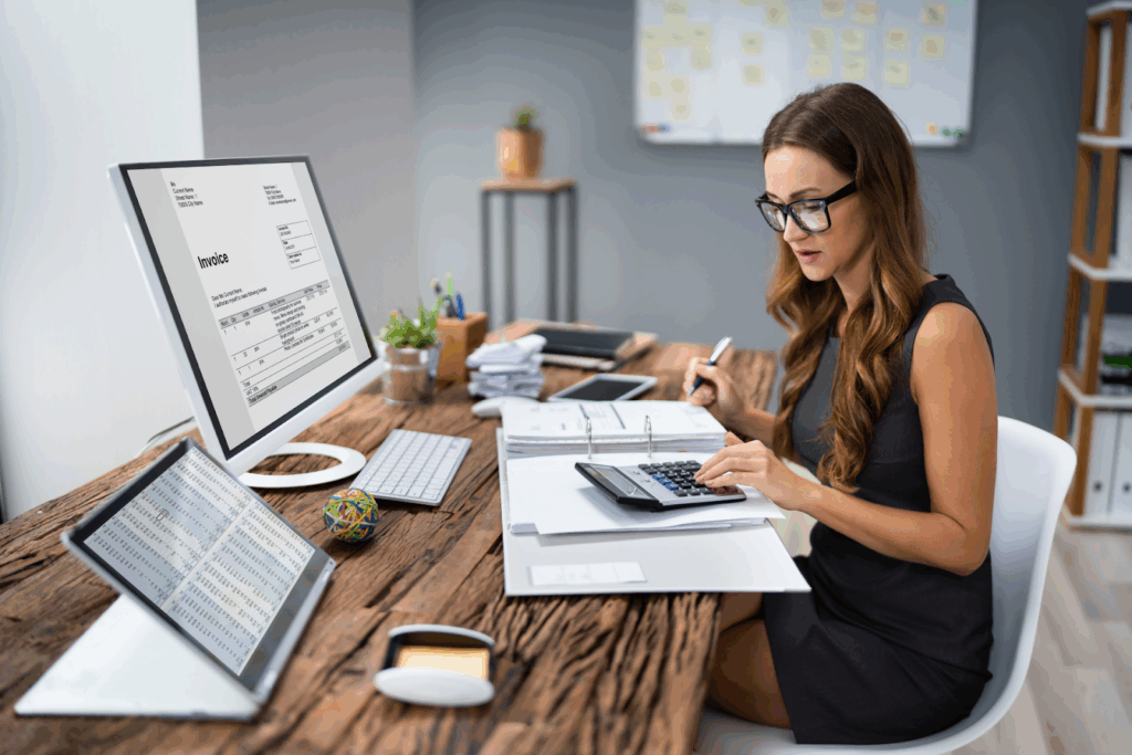 A woman in glasses uses a calculator while reviewing invoices and spreadsheets at her desk with dual monitors