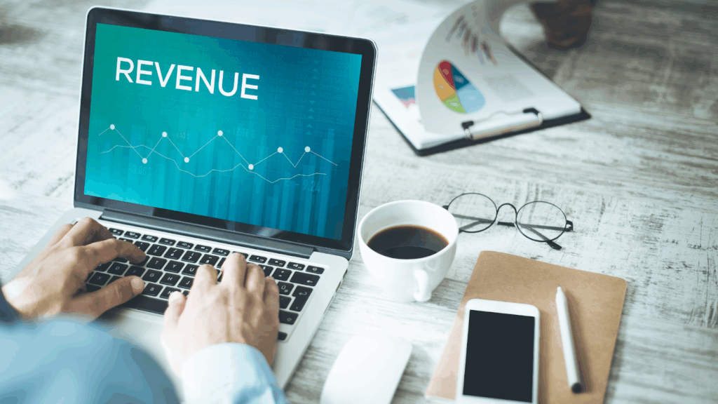 Person typing on a laptop with REVENUE displayed on screen next to coffee phone and financial charts