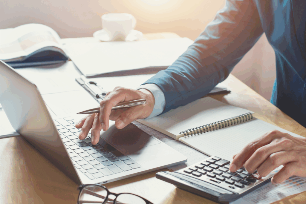 Person using a calculator and laptop at a desk with documents and a notebook