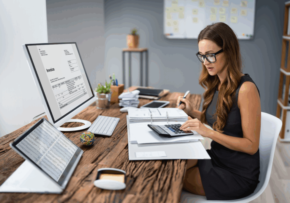 A woman in glasses uses a calculator while reviewing invoices and spreadsheets at her desk with dual monitors
