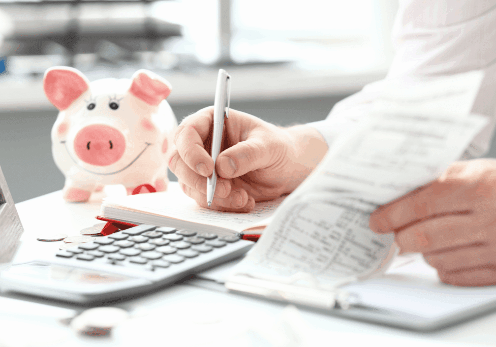 Person reviewing receipts and writing in a notebook with a piggy bank and calculator on the desk