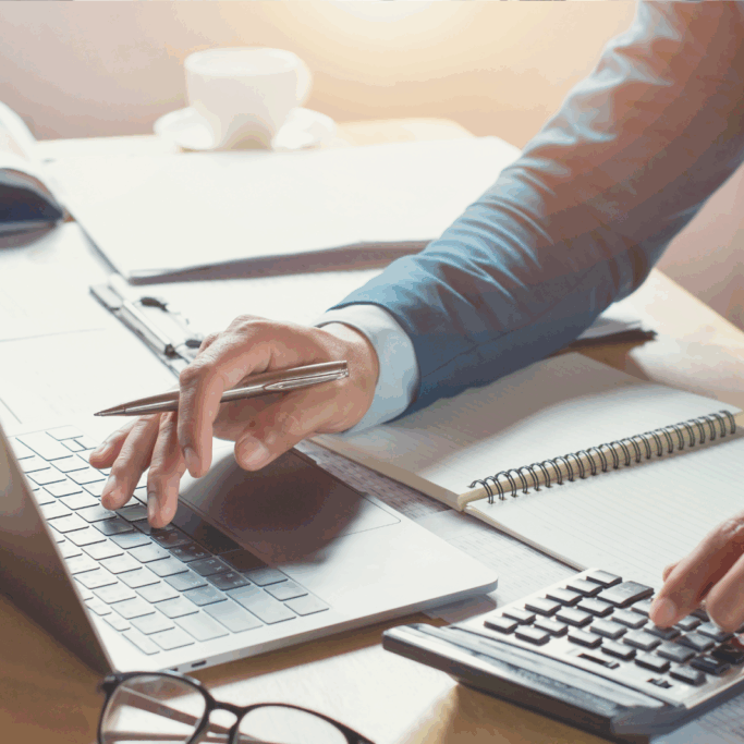 Person using a calculator and laptop at a desk with documents and a notebook