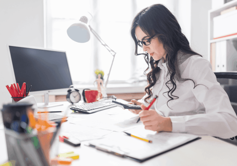 A focused woman in glasses working at her desk, using a calculator and writing notes with colorful stationery scattered around