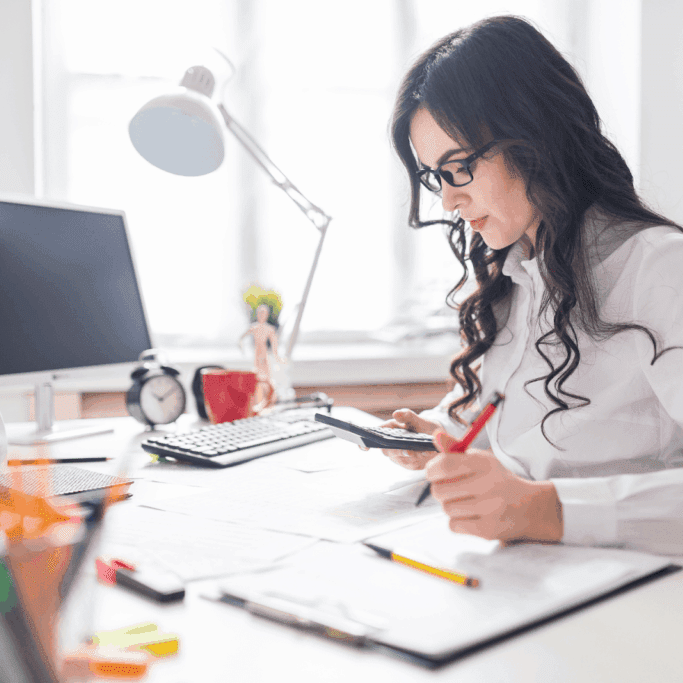 A focused woman in glasses working at her desk, using a calculator and writing notes with colorful stationery scattered around